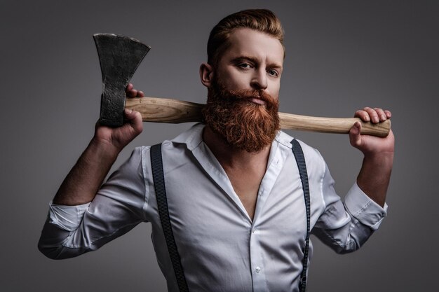 Brutal hombre barbudo maduro con barba pelirroja y bigote en tirantes retro sostiene hacha aislado en gris