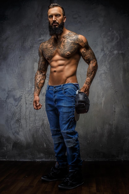 Brutal hombre tatuado sin camisa con barba en jeans azules. Posando sobre fondo gris.