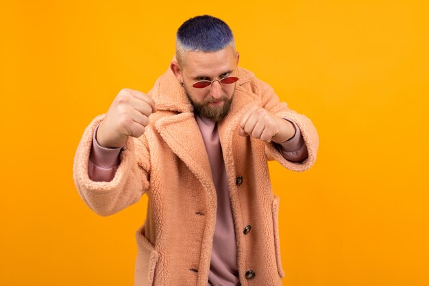 Brutaler kaukasischer Mann mit kurzen blauen Haaren und roten Gläsern in einem Pelzmantel, der auf Orange lokalisiert wird