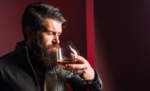 Brutaler Mann, der Whiskey schmeckt, starker Alkohol, bärtiger Typ in Lederjacke, der Brandy oder Cognac schnüffelt