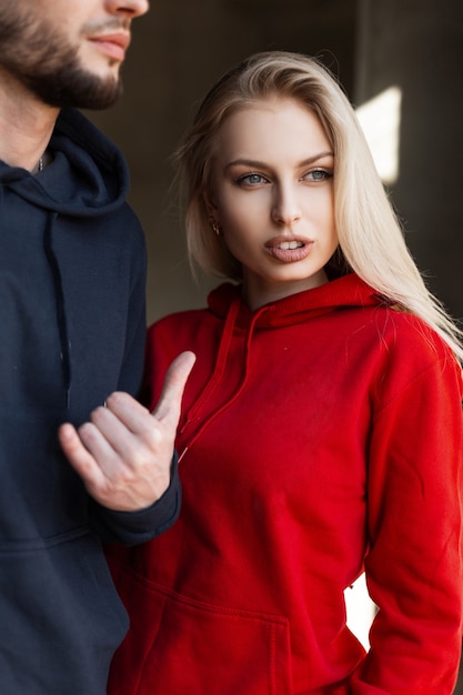 Brutaler Mann mit Bart in einem modischen Sweatshirt steht mit einer hübschen sexy jungen blonden Frau in einem roten trendigen roten Sweatshirt im Studio