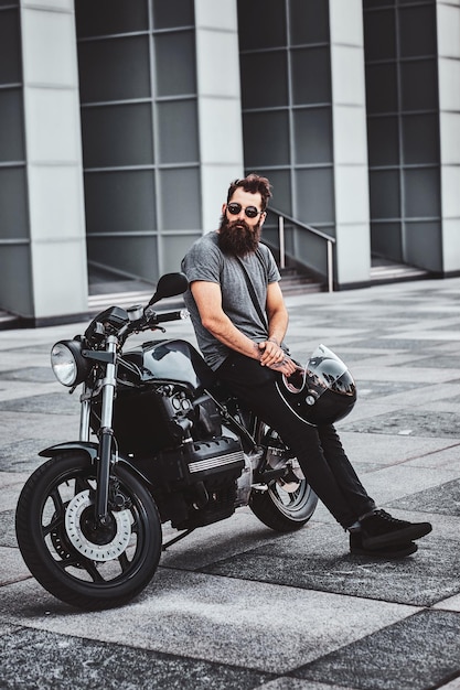 Brutaler tätowierter Biker mit Sonnenbrille ruht neben seinem Motorrad im Stadtzentrum.