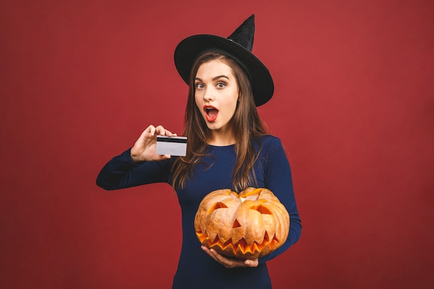 Bruxa de halloween com uma abóbora esculpida e cartão de crédito - isolado sobre fundo vermelho. jovem emocional em traje de halloween.