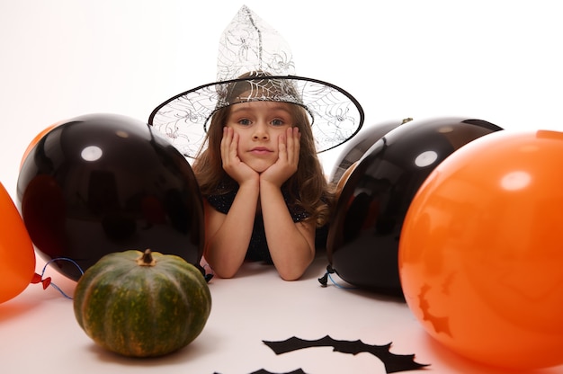 Bruxinha bonita com um chapéu de feiticeiro, deitada sobre um fundo branco com espaço de cópia ao lado de morcegos de feltro feitos à mão, abóbora e balões pretos laranja. Evento tradicional, conceito de festa de Halloween.
