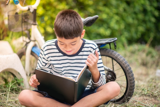 Buch des kleinen Jungen Lese, das mit Fahrrad im Park sitzt