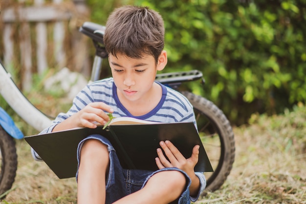 Buch des kleinen Jungen Lese, das mit Fahrrad im Park sitzt