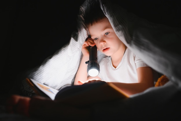 Buch lesen und Taschenlampe benutzen. Kleiner Junge in Freizeitkleidung, der sich abends in der Nähe des Zeltes hinlegt.