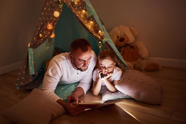 Buch lesen und Taschenlampe verwenden Vater und seine jungen Jungen verbringen Zeit zusammen drinnen in der Nähe des Zeltes
