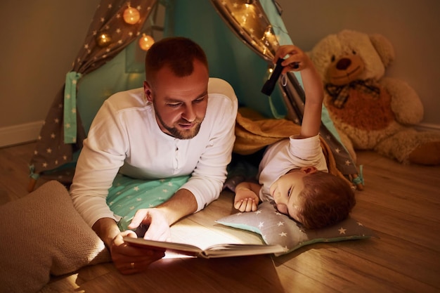 Buch lesen und Taschenlampe verwenden Vater und seine jungen Jungen verbringen Zeit zusammen drinnen in der Nähe des Zeltes