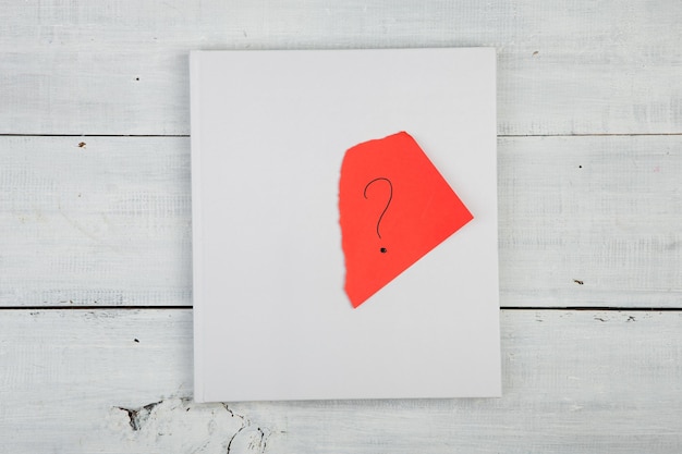 Buch und rotes Blatt mit einem Fragezeichen auf einem weißen Holzschreibtisch