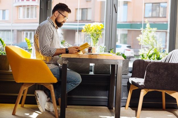 Buchliebhaber. Gut aussehender bärtiger Mann, der ein Buch in den Händen hält, während er in der Cafeteria liest