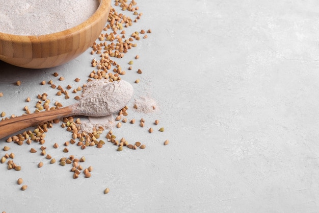 Buchweizenmehl in einer Holzschüssel und in einem Holzlöffel und rohes grünes Buchweizenkorn in einer Schüssel auf Grau