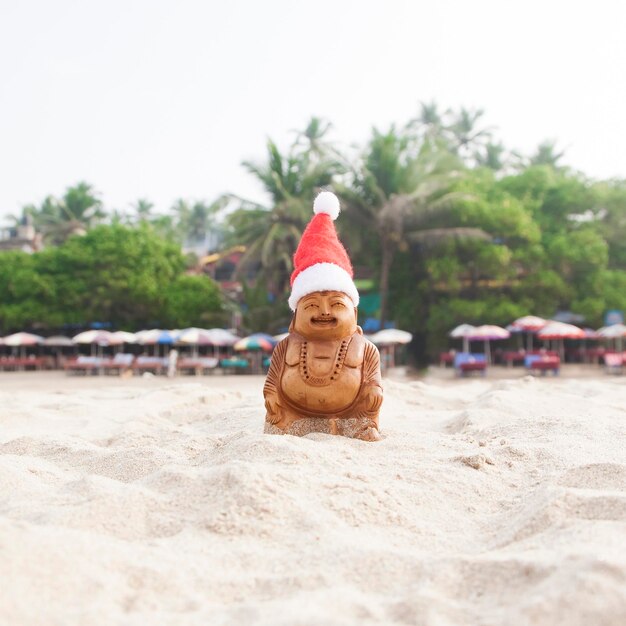 Budda-Statuette in der Weihnachtsmütze am Strand Tropischer Dschungel im Hintergrund