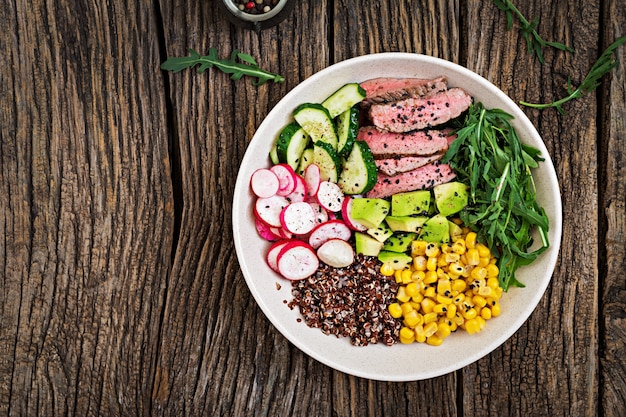 Buddha Bowl Lunch mit gegrilltem Rindersteak und Quinoa, Mais, Avocado, Gurke und Rucola auf Holztisch