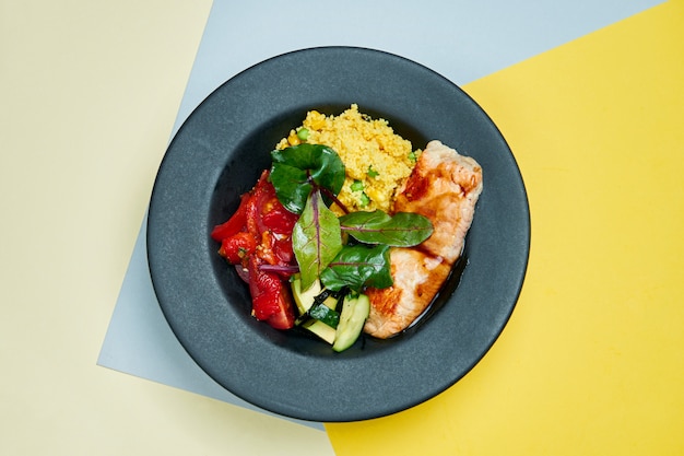 Buddha-Schale mit Lachsfilet, getrockneten Tomaten, Spinat, Gurke und Quinoa in einem schwarzen Teller auf einer farbigen Oberfläche Nahaufnahme. Suoer Essen