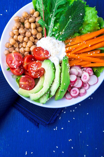 Buddha-Schüssel des strengen Vegetariers auf blauem hölzernem. Schüssel mit Karotten, Salat, Tomaten, Kirschen, Radieschen, Avocado und Kichererbsen. Vegetarisches, gesundes, entgiftendes Essen. Ansicht von oben.