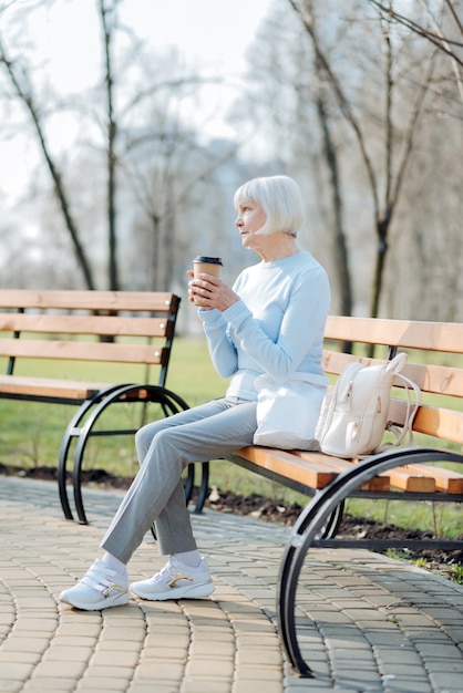 Buen café. Alegre mujer rubia bebiendo café mientras está sentado en el banco