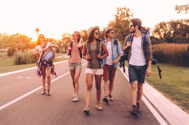 Buen día para hacer senderismo. Grupo de jóvenes con mochilas caminando juntos por la carretera y mirando felices