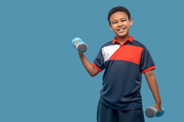 Buen estado físico. Alegre niño de piel oscura en ropa deportiva haciendo deporte con pesas de pie sobre fondo azul.