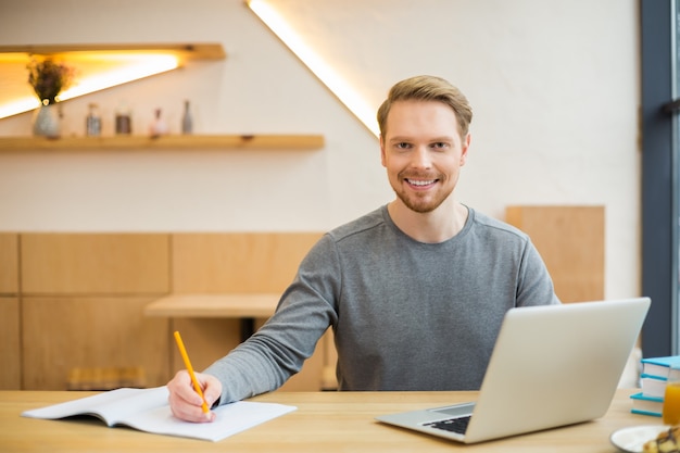 Buen hombre alegre positivo sonriendo y sosteniendo un lápiz mientras toma notas