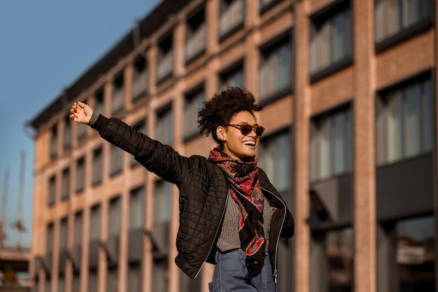 Buen humor. Una chica de piel oscura con gafas de sol caminando por la calle y sintiéndose alegre