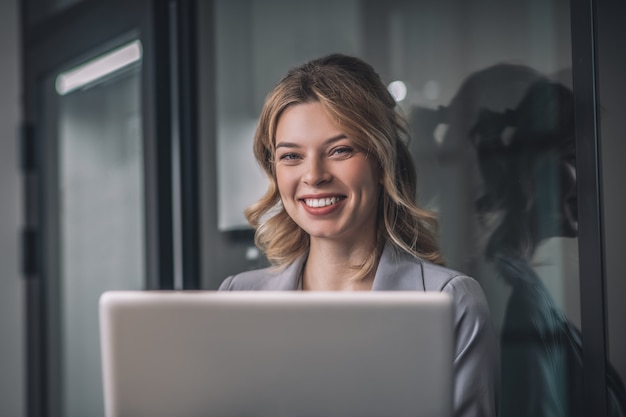 Buen humor. Feliz regocijo joven mujer de negocios en chaqueta gris con portátil de pie en el espacio de oficina