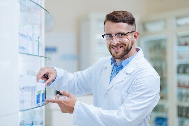 Buen humor. Hombre de cabello oscuro complacido manteniendo una sonrisa en su rostro mientras coloca la medicina en los estantes