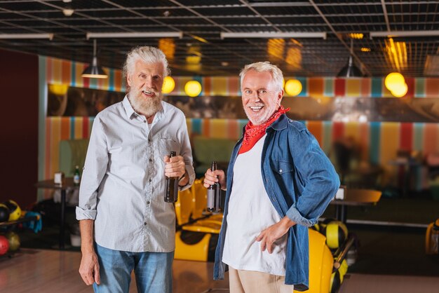 Buen humor. Hombres alegres positivos de pie bebiendo cerveza mientras se divierten en la bolera