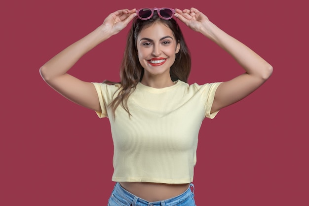 Buen humor. Mujer bonita joven sonriente con las manos cerca de la cabeza sosteniendo gafas de sol de buen humor