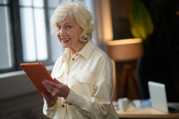 Buen humor. Sonriente anciana de pelo gris sosteniendo una tableta mirando a la cámara de pie en una habitación acogedora durante el día