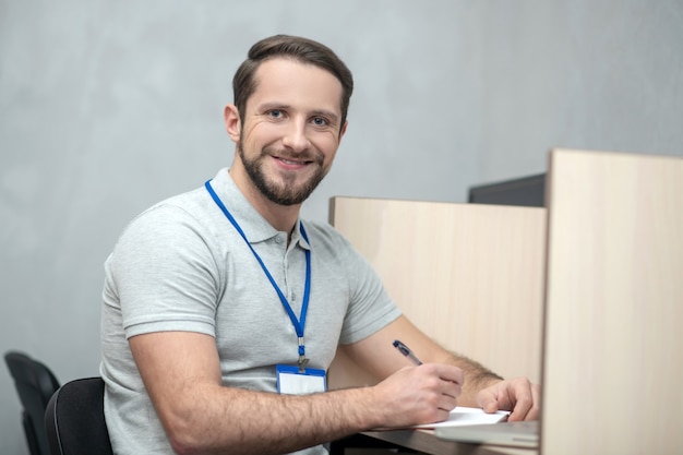 Buen humor. Sonriente joven adulto con placa sentado en la oficina trabajando de buen humor