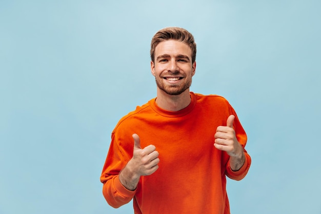 Buen joven de buen humor con barba de jengibre en sudadera fresca sonriendo mirando a la cámara y posando sobre fondo azul