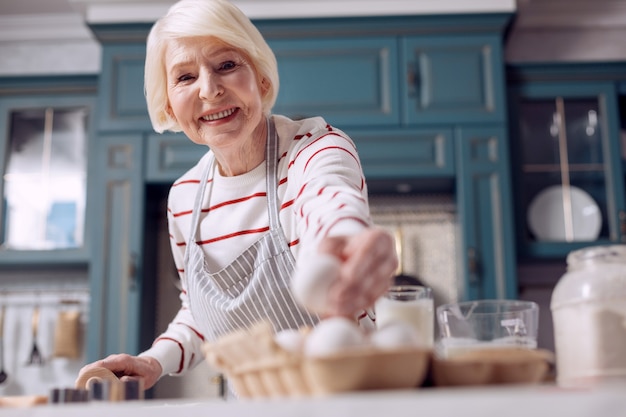 Buen panadero. La atención se centra en una agradable anciana que toma un huevo de un cartón de huevos y sonríe a la cámara mientras prepara la masa para un pastel.