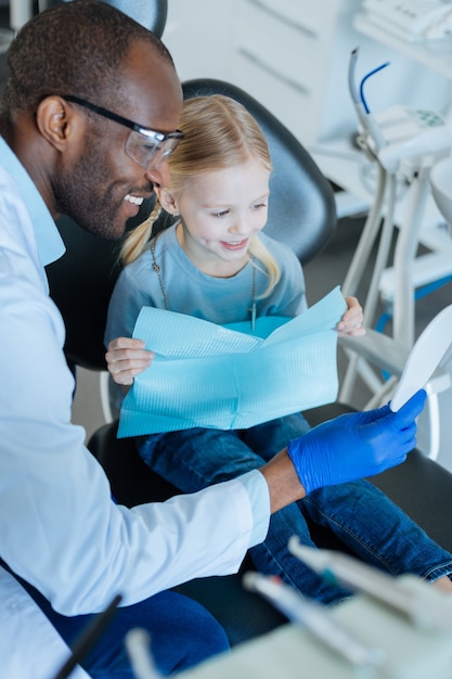 Buen resultado. niña linda sentada en la silla del dentista y mirándose en el espejo después del tratamiento mientras su agradable dentista lo sostiene para ella