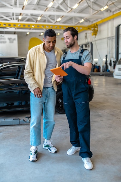 Foto buen servicio. joven mecánico barbudo hablando con confianza apuntando a la pantalla de la tableta y al cliente negro que parece interesado en el taller