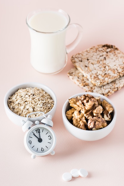 Buen sueño. Alimentos para dormir bien: leche, nueces, pan tostado, avena y despertador sobre fondo rosa. Vista vertical