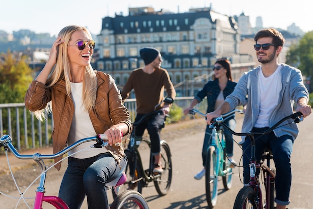 Buen tiempo con amigos. Hermosa joven sonriente montando bicicleta y mirando a otro lado mientras sus amigos cabalgan en el fondo