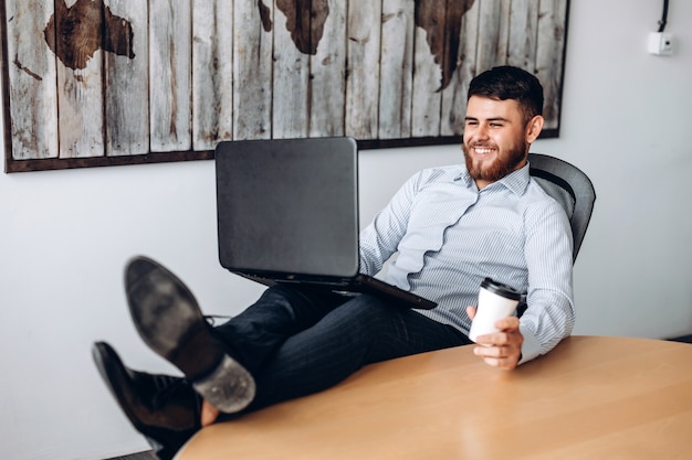 Buen tipo con barba sentado en una mesa, poniendo los pies sobre la mesa, tomando café y trabajando en una computadora