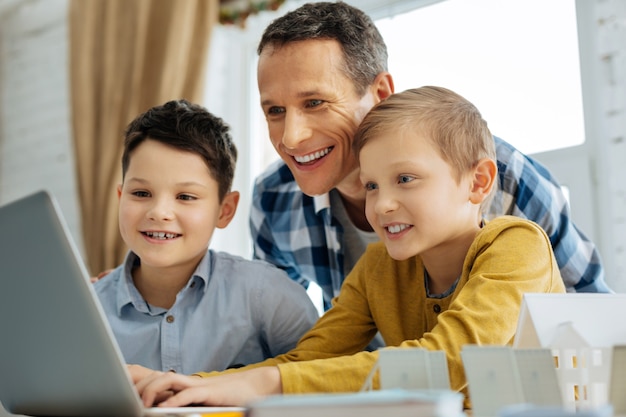 Buen trabajo. Padre joven optimista viendo la presentación del proyecto de sus hijos en la computadora portátil relacionado con las fuentes alternativas de energía y sonriendo, orgulloso de ellos