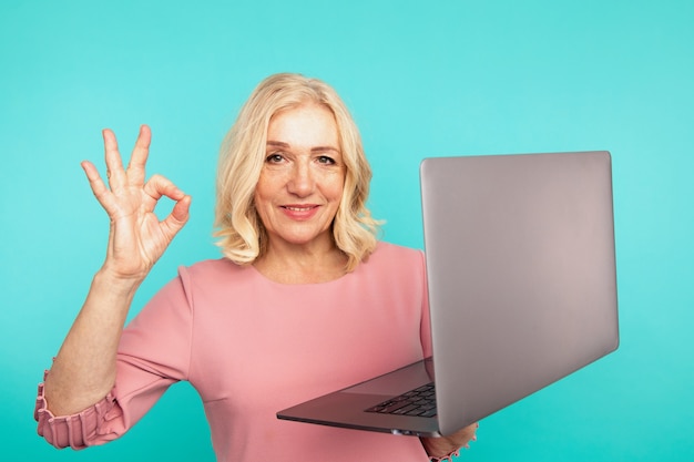 Buen trabajo portátil. Mujer mostrando signo ok con computadora en la habitación azul.