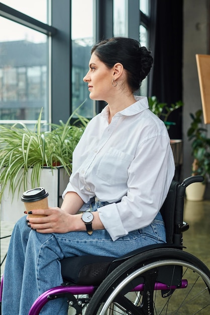 Foto buena apariencia mujer de negocios alegre en ropa casual en silla de ruedas sosteniendo café mientras está en la oficina