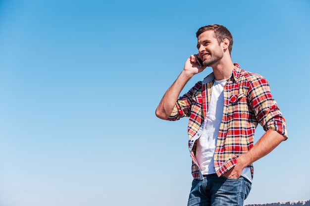 Buena charla con un amigo. Joven alegre hablando por teléfono móvil y sonriendo mientras está de pie al aire libre con el cielo azul como fondo