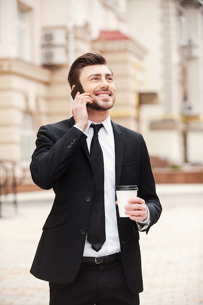 Buena charla de negocios. Apuesto joven en ropa formal hablando por teléfono móvil y sonriendo mientras está de pie al aire libre