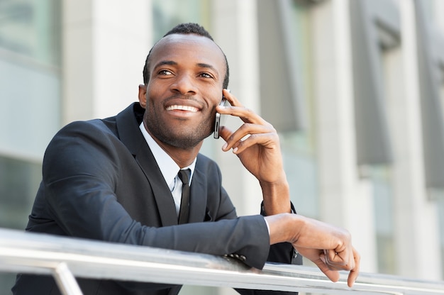 Buena charla de negocios. Feliz joven africano en ropa formal hablando por teléfono móvil y sonriendo mientras está de pie al aire libre