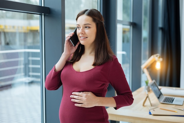 Buena conversacion. Joven mujer embarazada alegre hablando por teléfono móvil mientras expresa alegría y toca su estómago