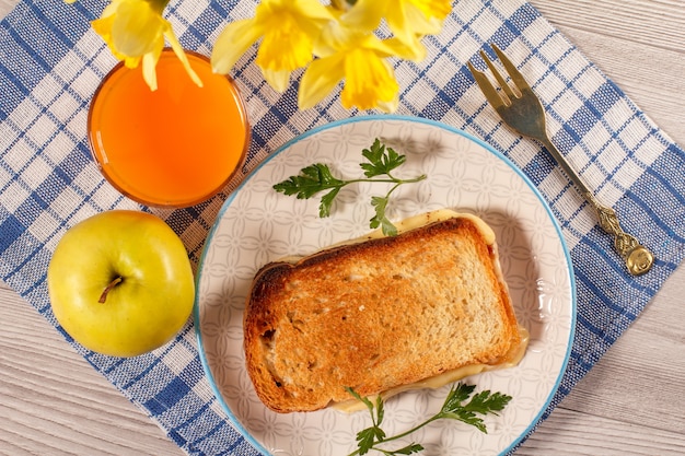 Buena y deliciosa comida y bebidas para el desayuno. Tostadas con mantequilla y queso en un plato blanco con tenedor, manzana, ramo de narcisos amarillos y vaso de jugo de naranja en una servilleta de cocina. Vista superior