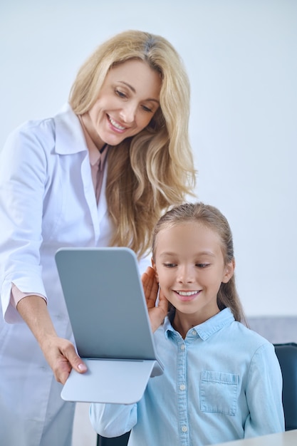 Buena elección. Brillante niña de edad escolar primaria complacida probándose un audífono y una amigable doctora sosteniendo un espejo