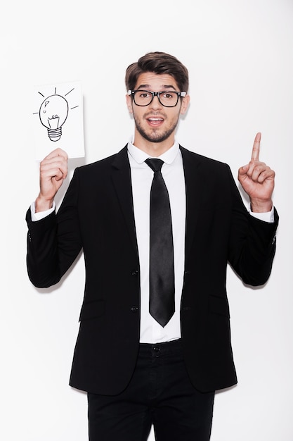 Foto ¡buena idea! hombre joven emocionado en ropa formal apuntando hacia arriba y sosteniendo la imagen de la lámpara eléctrica mientras está de pie contra el fondo blanco.