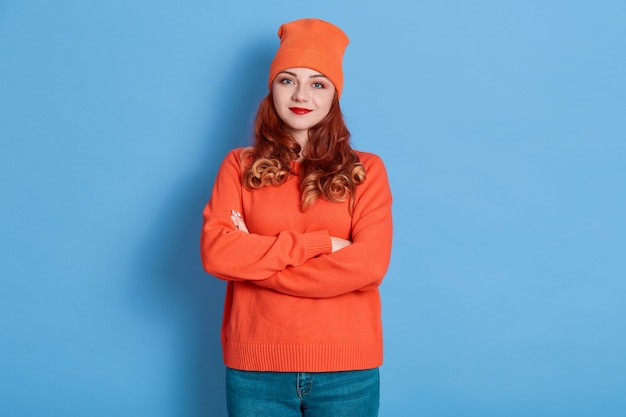 Buena mujer europea joven que está tranquila, mantiene los brazos cruzados, viste un suéter y gorra de punto naranja