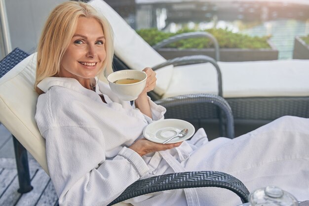 Buena mujer feliz vistiendo bata blanca descansando con una taza de té en el exterior